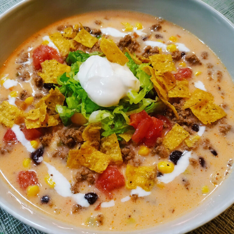 A close-up of a bowl of creamy taco soup topped with sour cream, shredded cheese, cilantro, avocado, and tortilla chips, showing ground beef, beans, and corn in a rich broth.