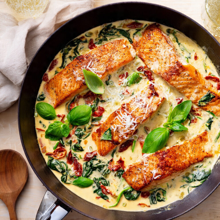 A cast iron skillet with creamy Tuscan salmon, showing pan-seared salmon fillets in a rich sauce with sun-dried tomatoes, spinach, and a garnish of Parmesan cheese and fresh basil.