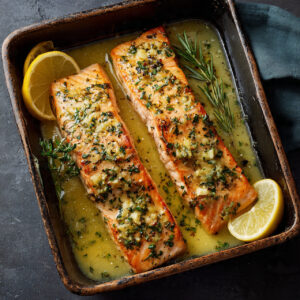 Overhead view of baked salmon fillets in a ceramic dish, covered in a glossy garlic butter sauce with herbs and lemon wedges.