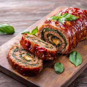 A sliced stuffed Italian meatloaf on a wooden board, showing a spiral of cheese, spinach, and sun-dried tomatoes.